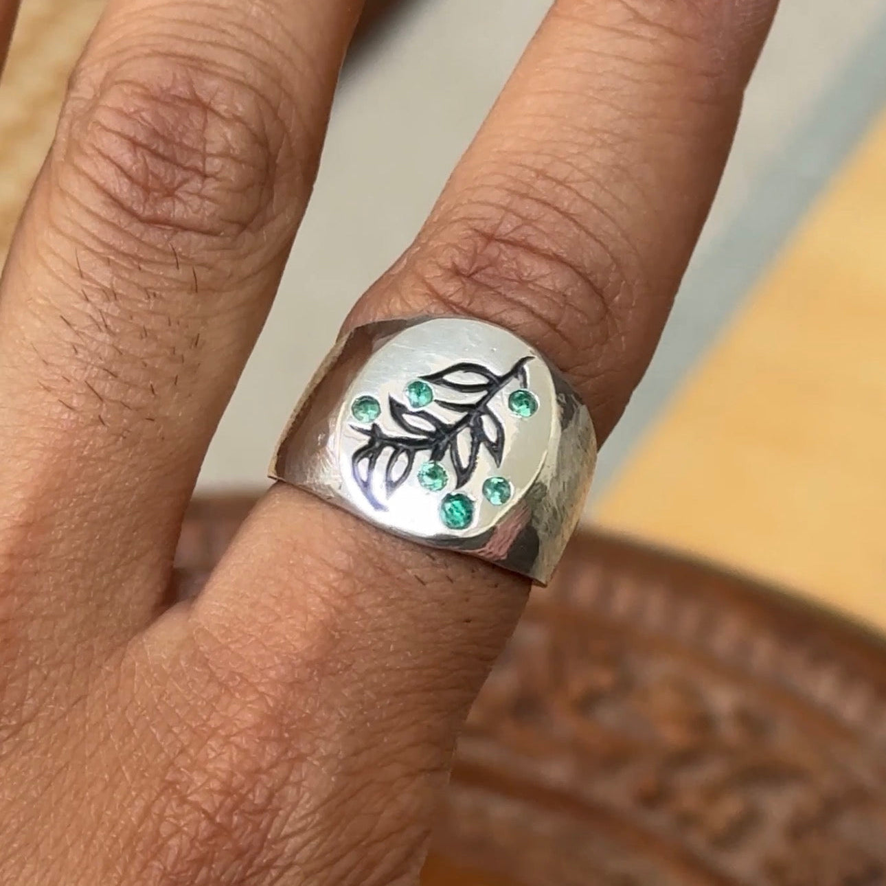 A close-up of a hand wearing a wide sterling silver ring with an engraved leaf design and small green stone accents.