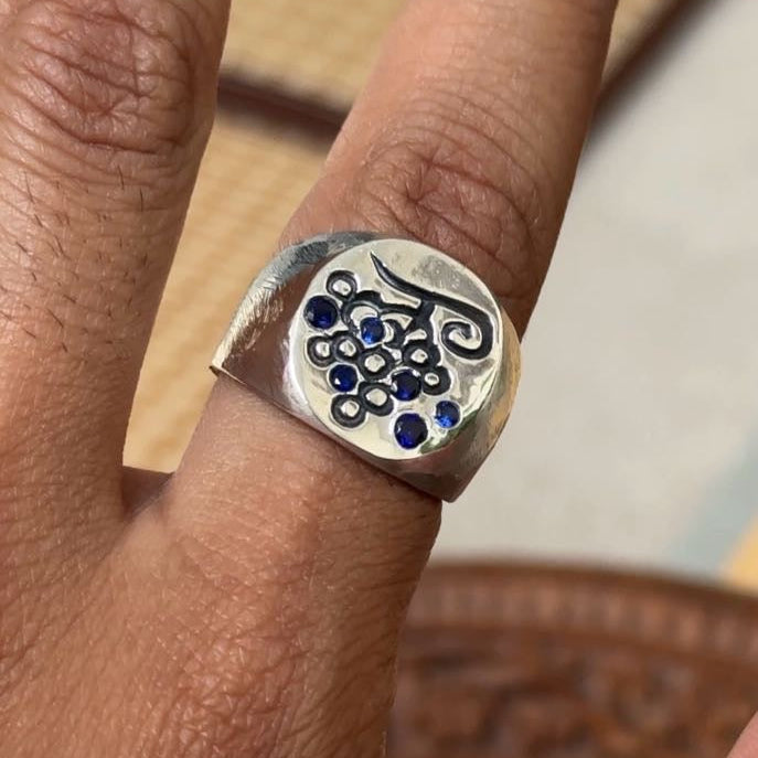 A silver ring with a stylized black script design and small blue gemstone accents, shown worn on a finger.