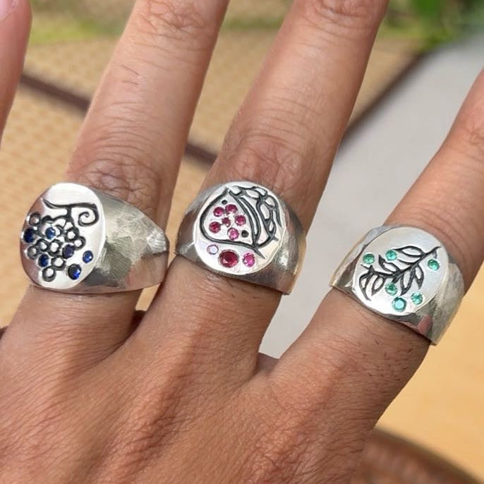 Three silver signet-style rings with engraved floral and fruit motifs, worn on a hand. The rings are accented with small blue, pink, and green stones.