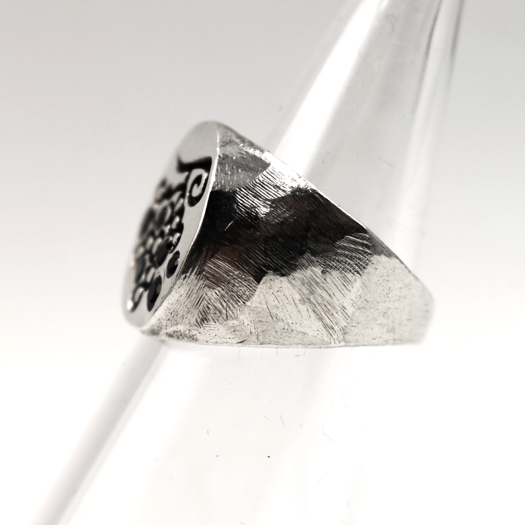 A silver signet-style ring with a hammered finish and a black engraved floral or scroll motif on one side, displayed on a clear plastic cone against a white background.