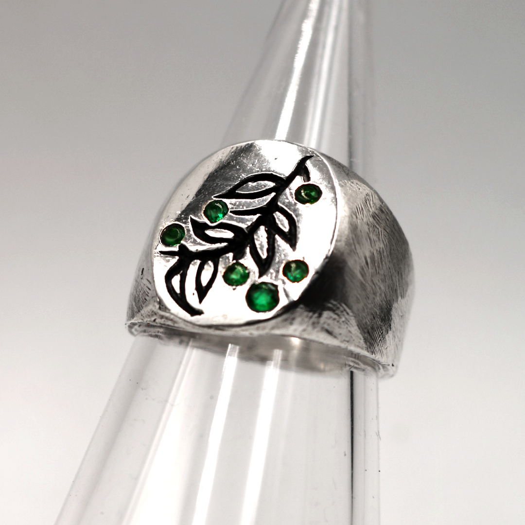 A sterling silver signet ring with a textured leaf design and small green gemstone accents, displayed on a clear plastic ring cone against a neutral gray background.