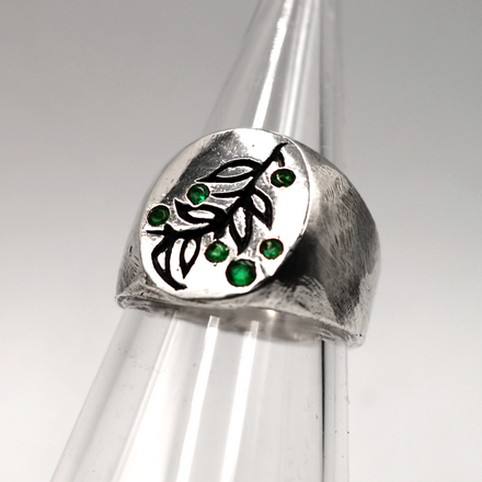 A sterling silver signet ring with a textured leaf design and small green gemstone accents, displayed on a clear plastic ring cone against a neutral gray background.