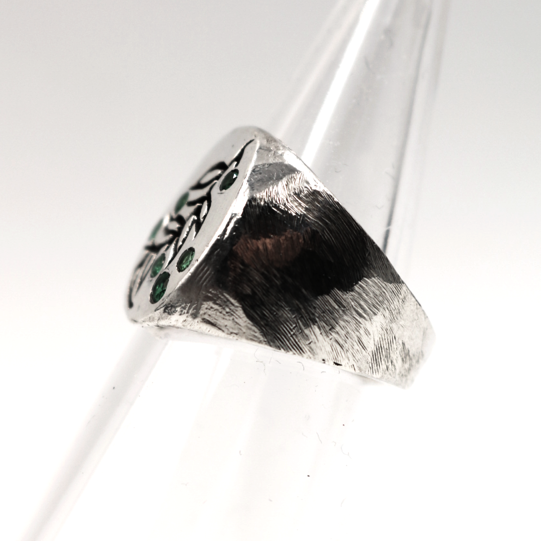 A sterling silver signet ring with a textured band and a flat face featuring a black engraved leaf pattern and small green stone accents, displayed on a clear plastic ring cone against a white background.