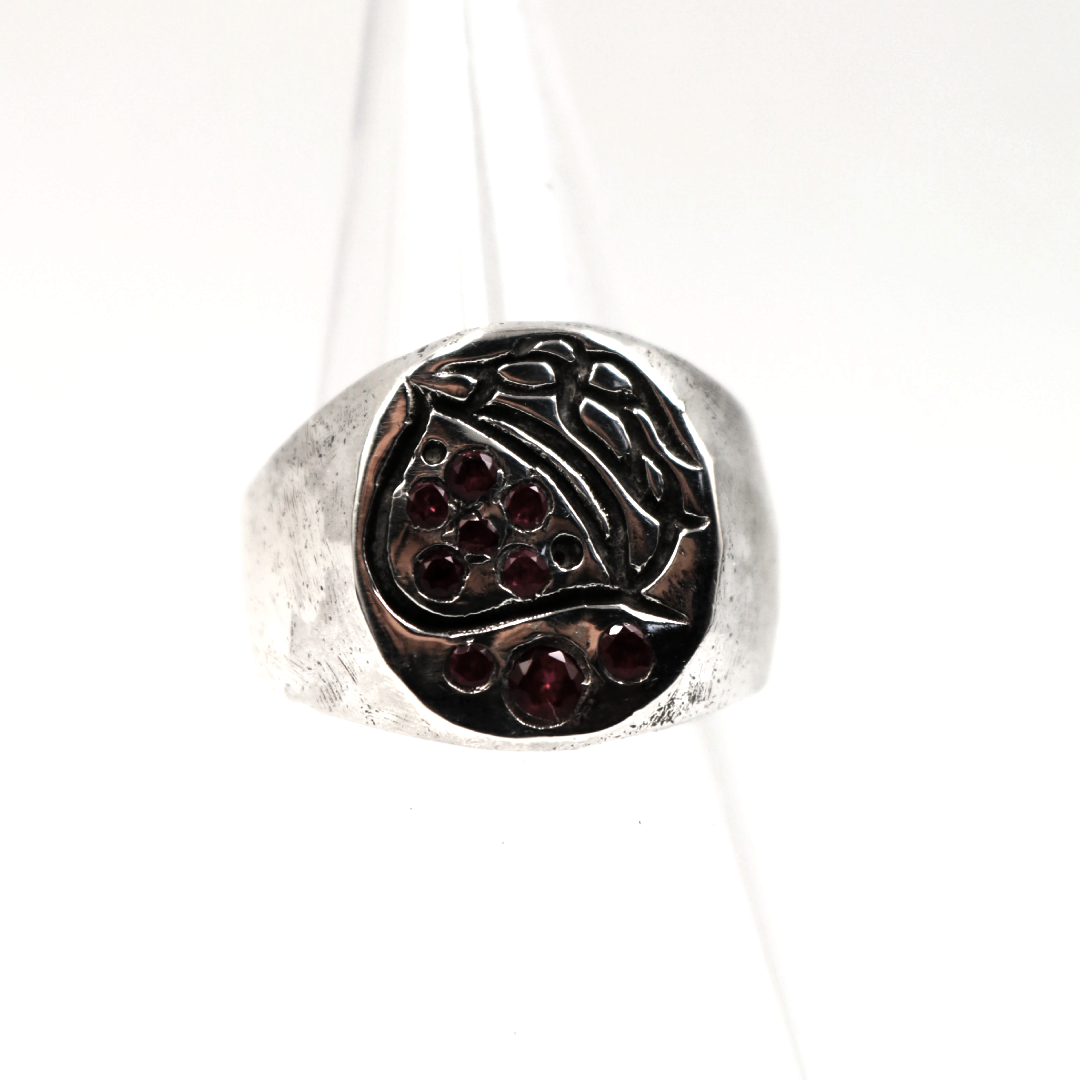 A silver signet ring with a dark, textured circular face featuring small red gemstone accents, displayed on a clear plastic ring stand against a white background.