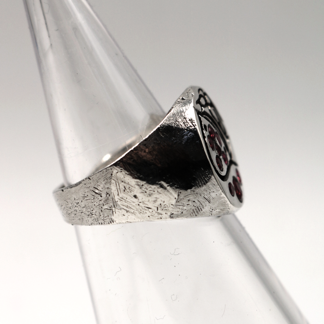 A silver signet ring with a textured band and a dark, patterned face, displayed on a clear plastic cone stand against a light gray background.